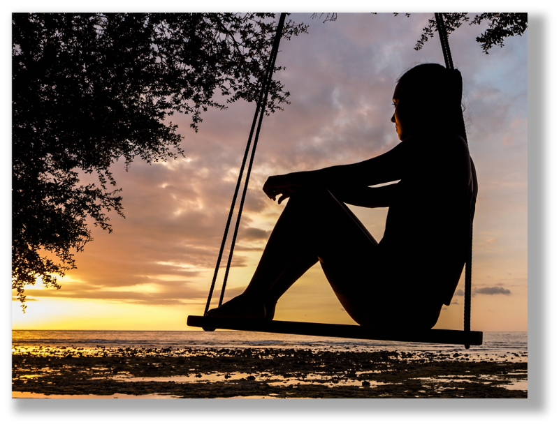 A woman sits on a swing, gazing out across a beach to a beautiful sunset.